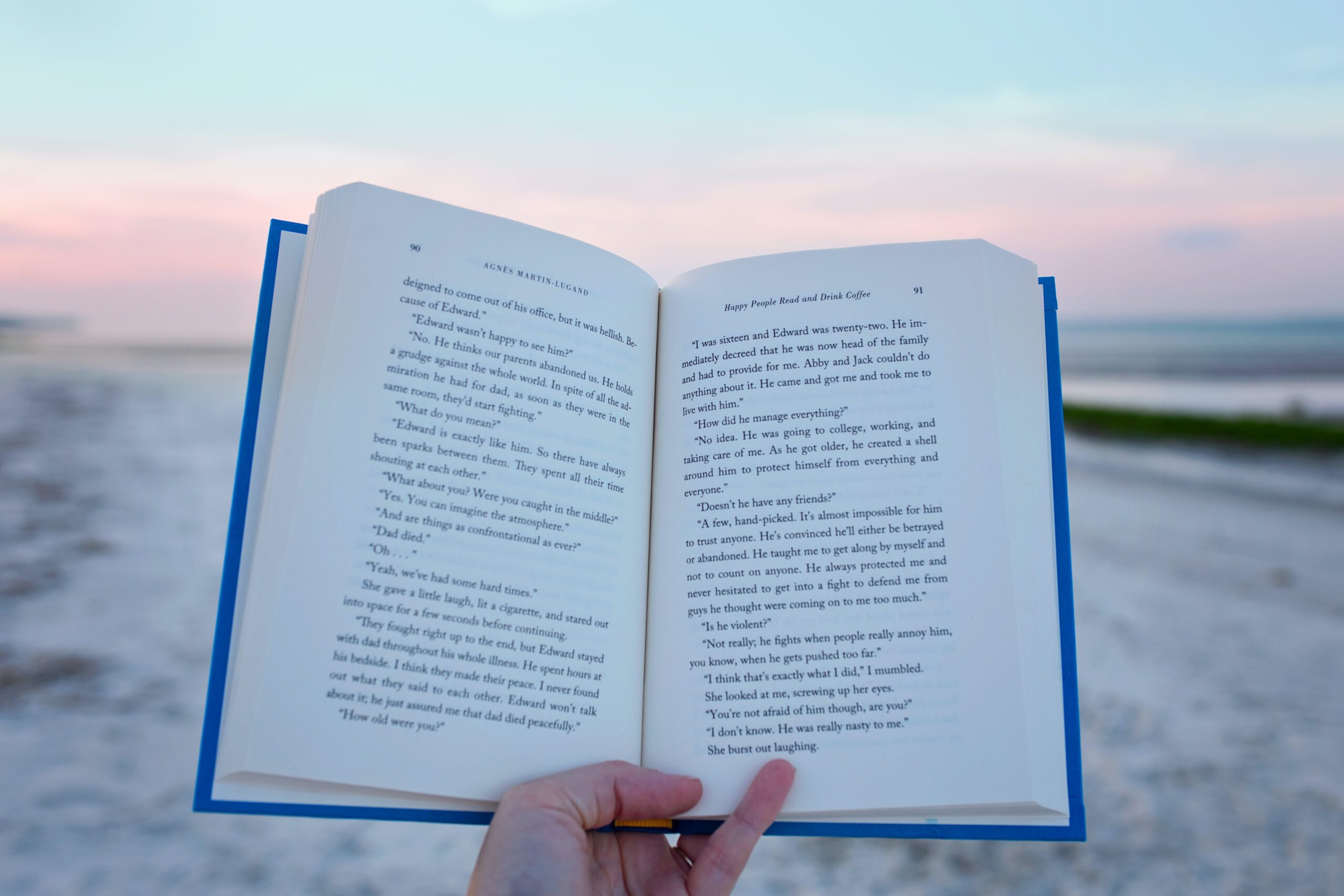 An image of an open book being held by a hand with the beach being a backdrop